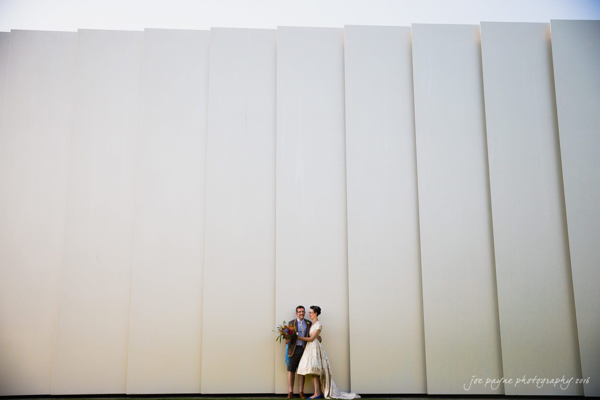 nc museum of art wedding photography - jade & frank A bride and groom stand closely together, holding a bouquet in front of a large, modern beige wall with vertical lines. The couple is smiling, and the groom is wearing a gray suit while the bride is in a white dress.