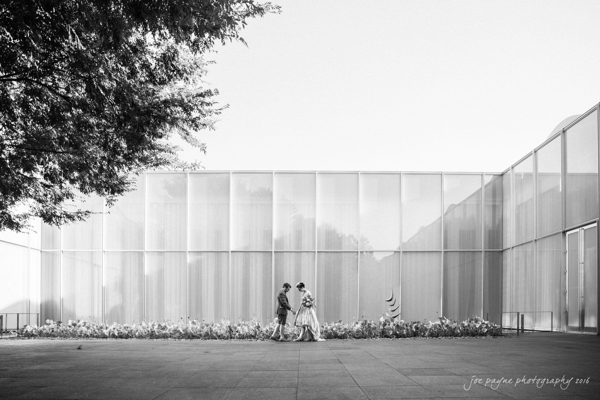 nc museum of art wedding photography - jade & frank A black and white photo of a couple walking hand in hand past a modern building with glass walls. They are surrounded by a landscaped flower bed, and a large tree casts a shadow on the left.