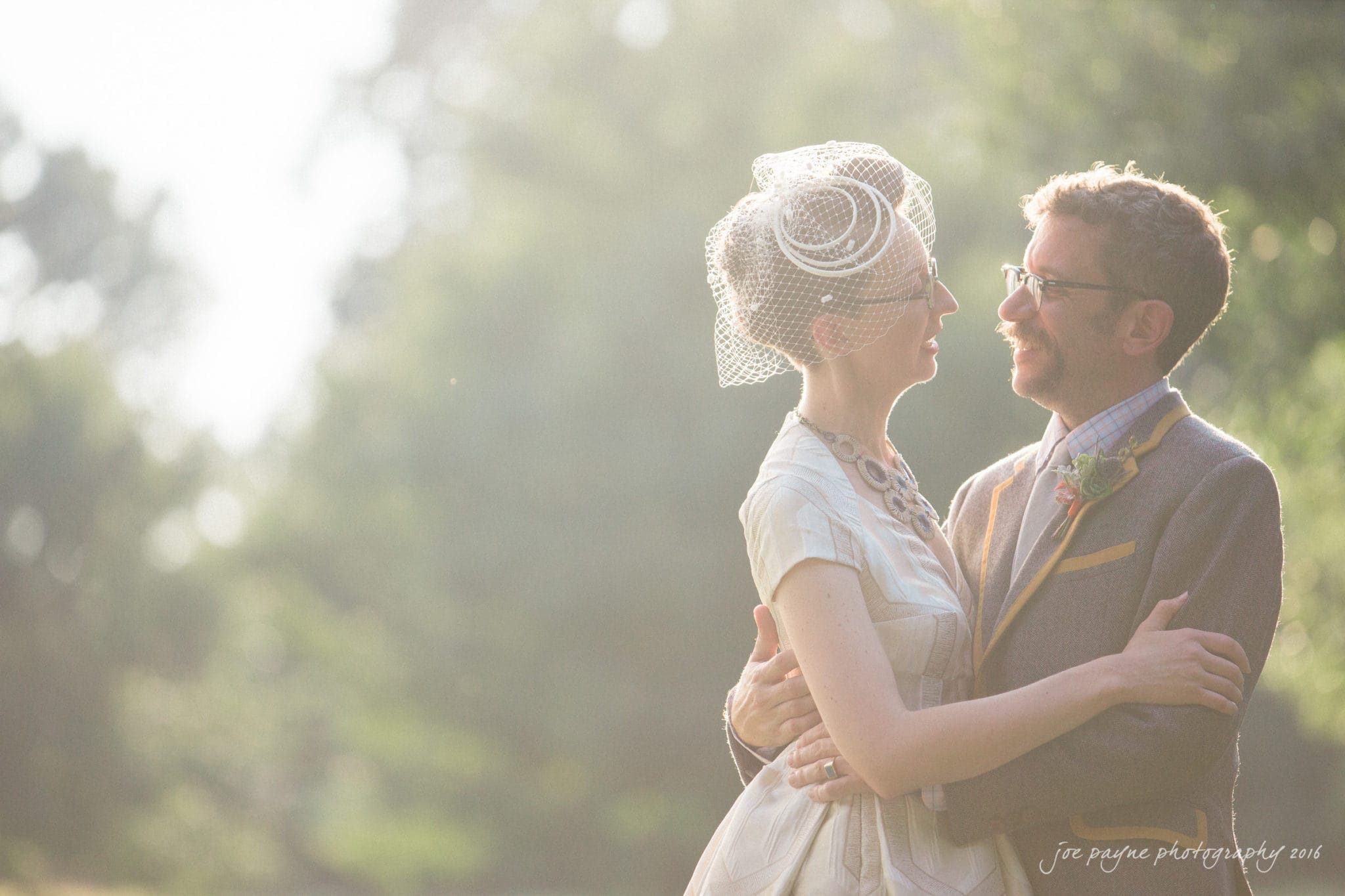 nc museum of art wedding photography - jade & frank A couple stands embraced in a sunlit outdoor setting. The woman wears a vintage-style dress and birdcage veil, while the man dons a suit and glasses. They gaze at each other lovingly, surrounded by greenery.