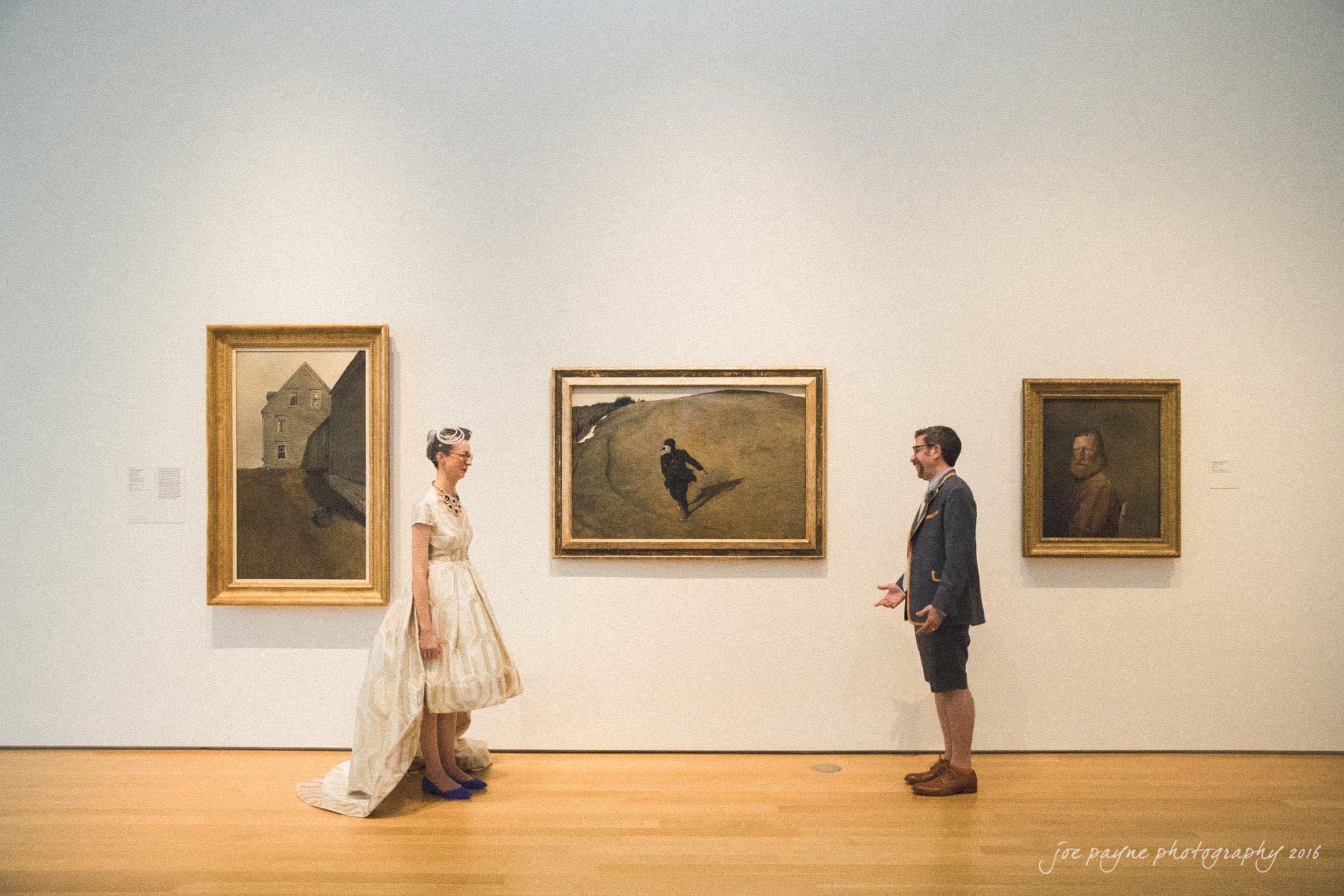 nc museum of art wedding photography - jade & frank A bride in a white dress and a groom in casual attire stand facing each other in an art gallery. Three paintings hang on the wall behind them, with a wooden floor beneath. The setting is bright and minimalist.