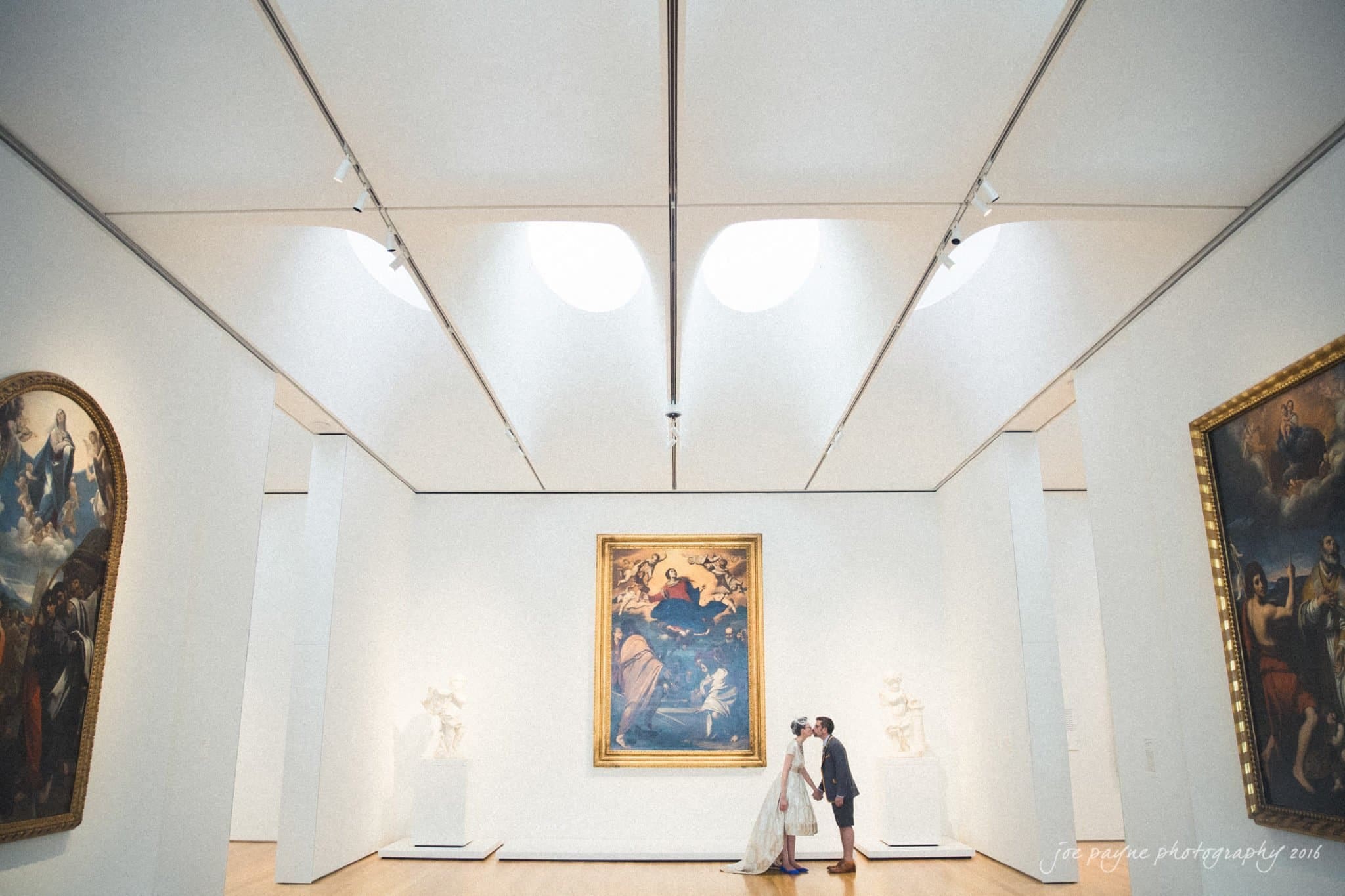 nc museum of art wedding photography - jade & frank A couple in wedding attire stands in the center of a bright gallery room, surrounded by large paintings and sculptures. The ceiling has a unique triangular pattern, and soft lighting illuminates the art displays.