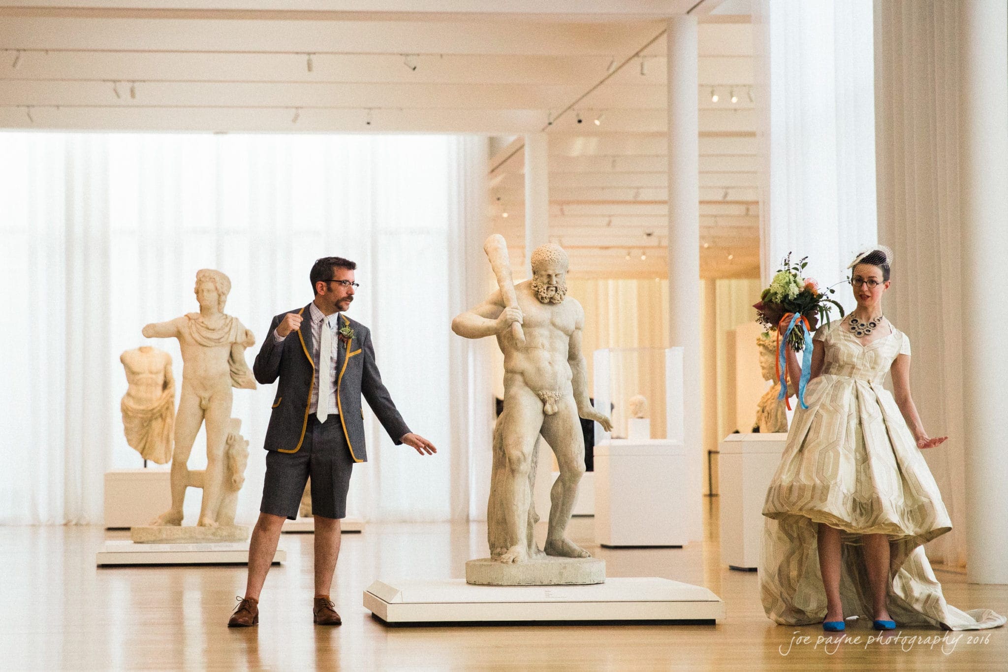 nc museum of art wedding photography - jade & frank A man and a woman are playfully posing in a bright gallery filled with classical statues. The man wears a suit with shorts, while the woman wears a whimsical dress and holds a bouquet. Both appear to be enjoying themselves in the artistic setting.