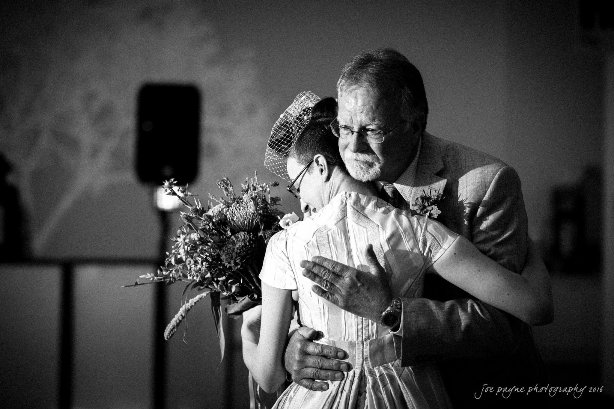 nc museum of art wedding photography - jade & frank A black and white photo of a man and woman hugging closely. The woman holds a bouquet of flowers. They appear emotional and are standing in a softly lit room.