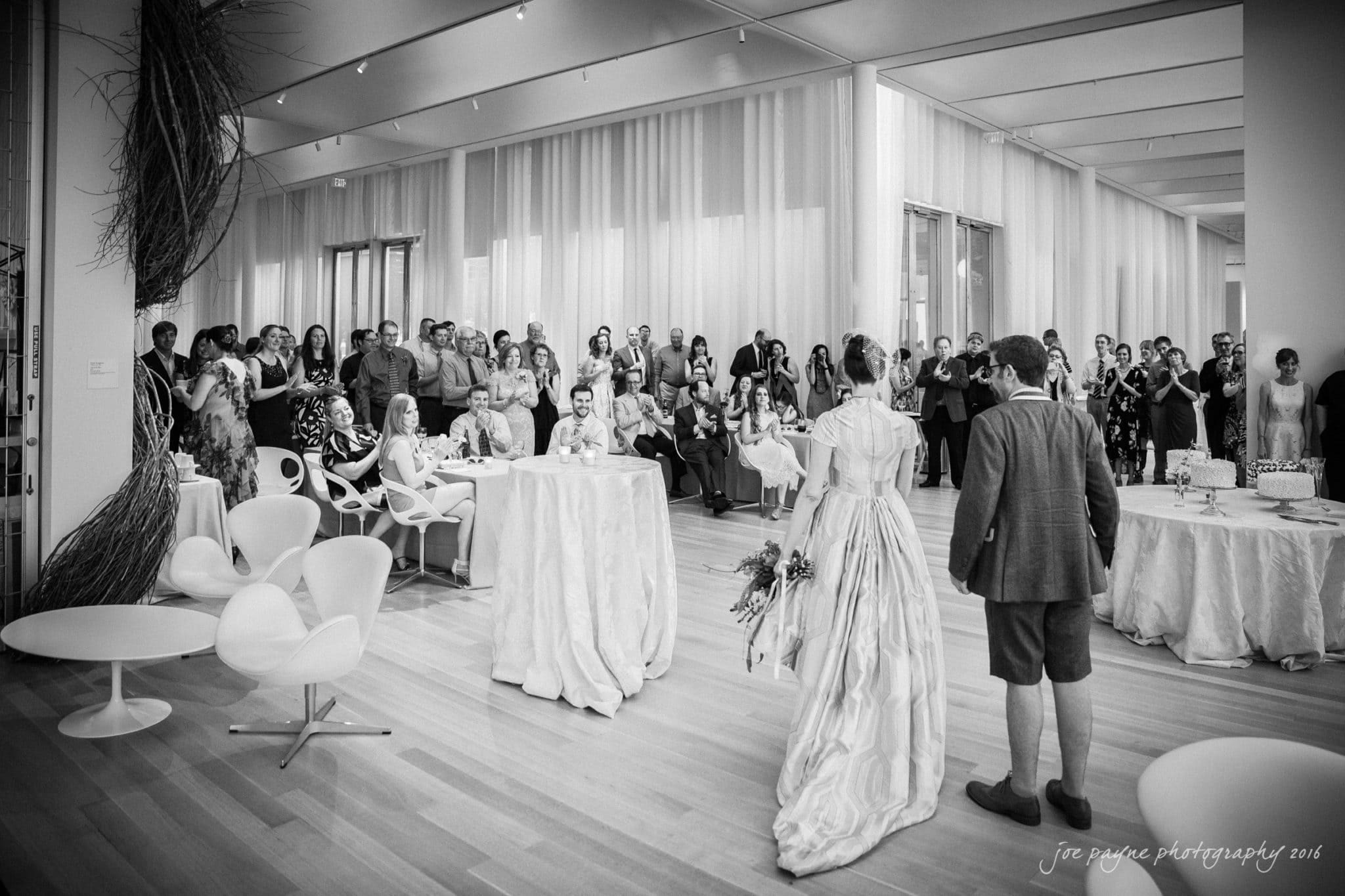 nc museum of art wedding photography - jade & frank A black and white photo of a bride and groom entering a modern reception hall filled with guests. The bride holds a bouquet, and the decor is elegant with draped tables and contemporary chairs. Guests are gathered around, watching the couple.