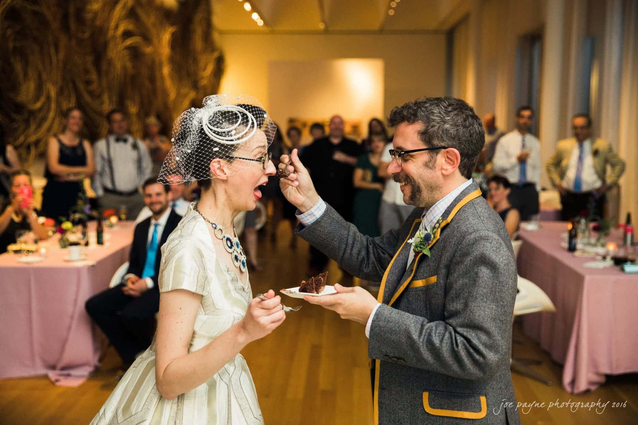 nc museum of art wedding photography - jade & frank A bride and groom laughing as the groom playfully offers her a bite of cake. They stand in a room with guests seated at tables in the background. The bride wears a vintage-style dress and veil, while the groom is in a gray suit with yellow accents.