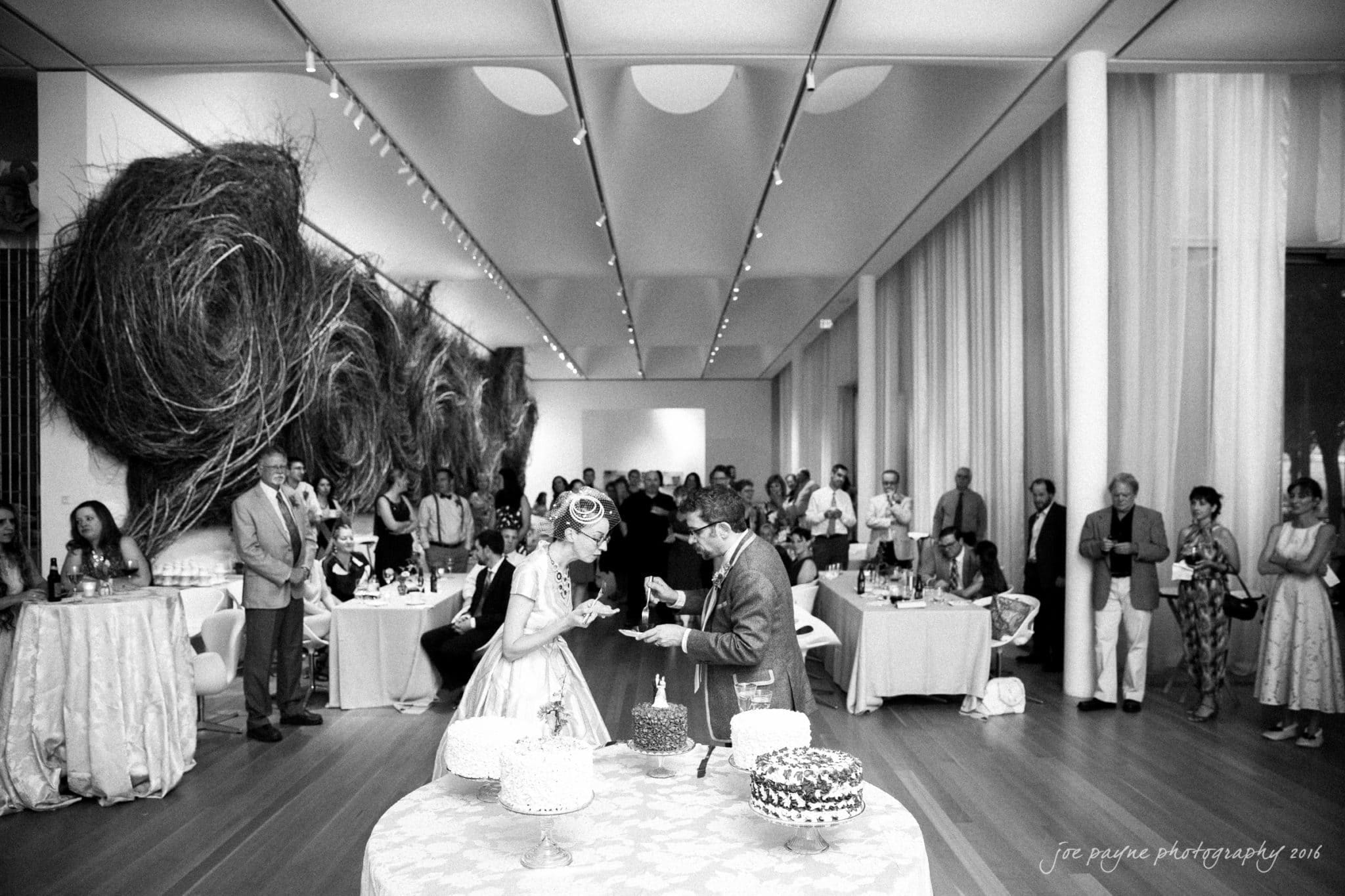 nc museum of art wedding photography - jade & frank A couple is cutting cake at their wedding reception in a modern, elegant venue. Guests stand and watch, surrounded by art and natural light from large windows. Several cakes are displayed on a table in the foreground.