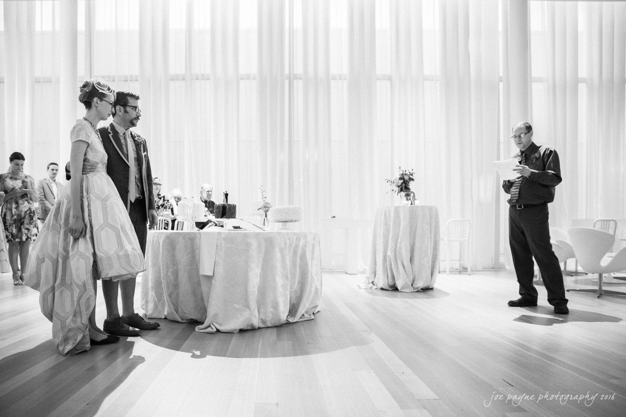nc museum of art wedding photography - jade & frank A bride and groom stand together, listening to a man giving a speech in a brightly lit room with large windows. A table with a cake and flowers is nearby. Guests are visible in the background.