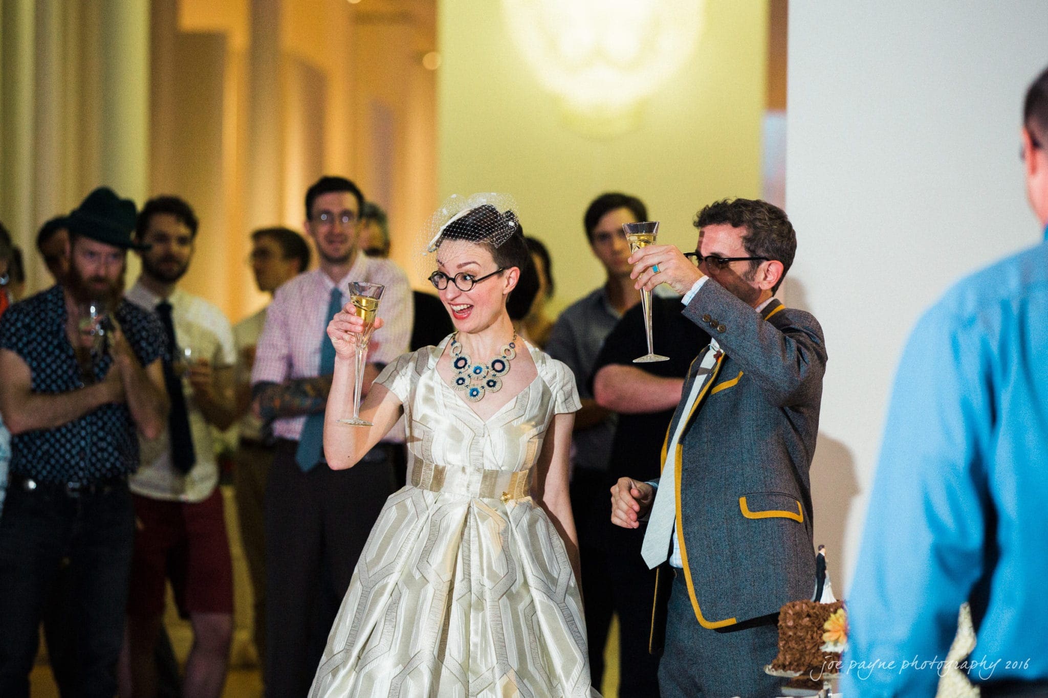 nc museum of art wedding photography - jade & frank A couple dressed in vintage-style attire raises their glasses in a toast at a gathering. The woman wears a white dress and veil, and the man dons a suit with a yellow trim. Guests in casual attire applaud in the background.