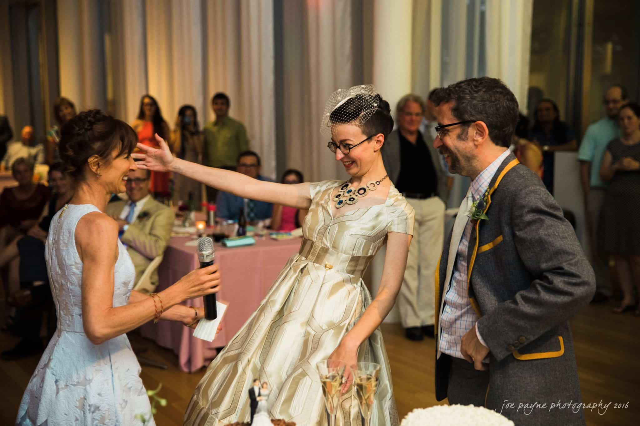 nc museum of art wedding photography - jade & frank A bride in a white dress and veil joyfully hugs a woman with a microphone, while a man in glasses and a suit stands smiling nearby. A crowd watches in the background, and a cake with a bride and groom topper is in the foreground.