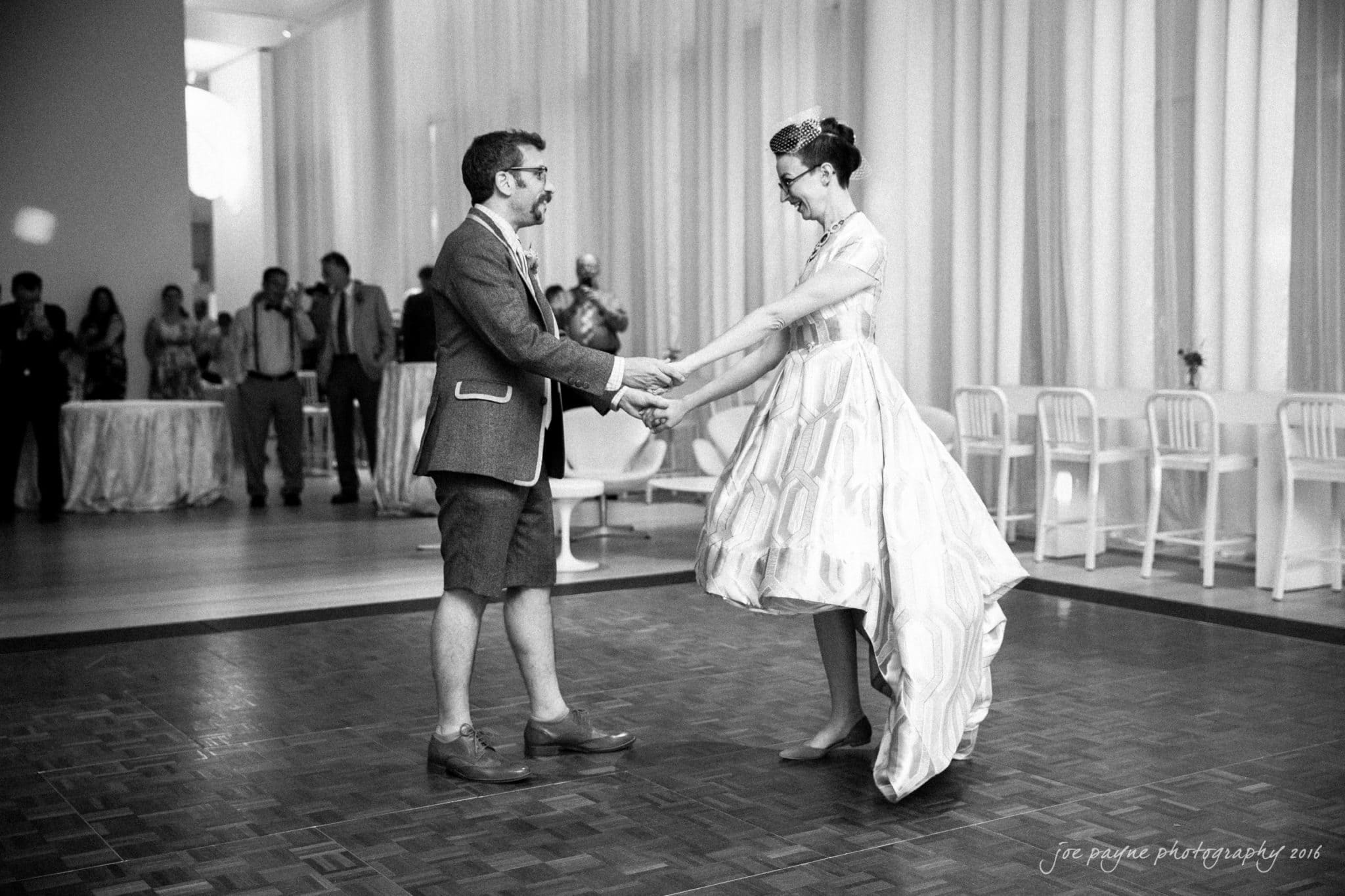 nc museum of art wedding photography - jade & frank A couple dances together on a wooden floor in a formal setting. The woman wears a vintage-style dress, while the man sports a suit with shorts. Onlookers stand in the background, watching and clapping. The atmosphere is elegant and festive.