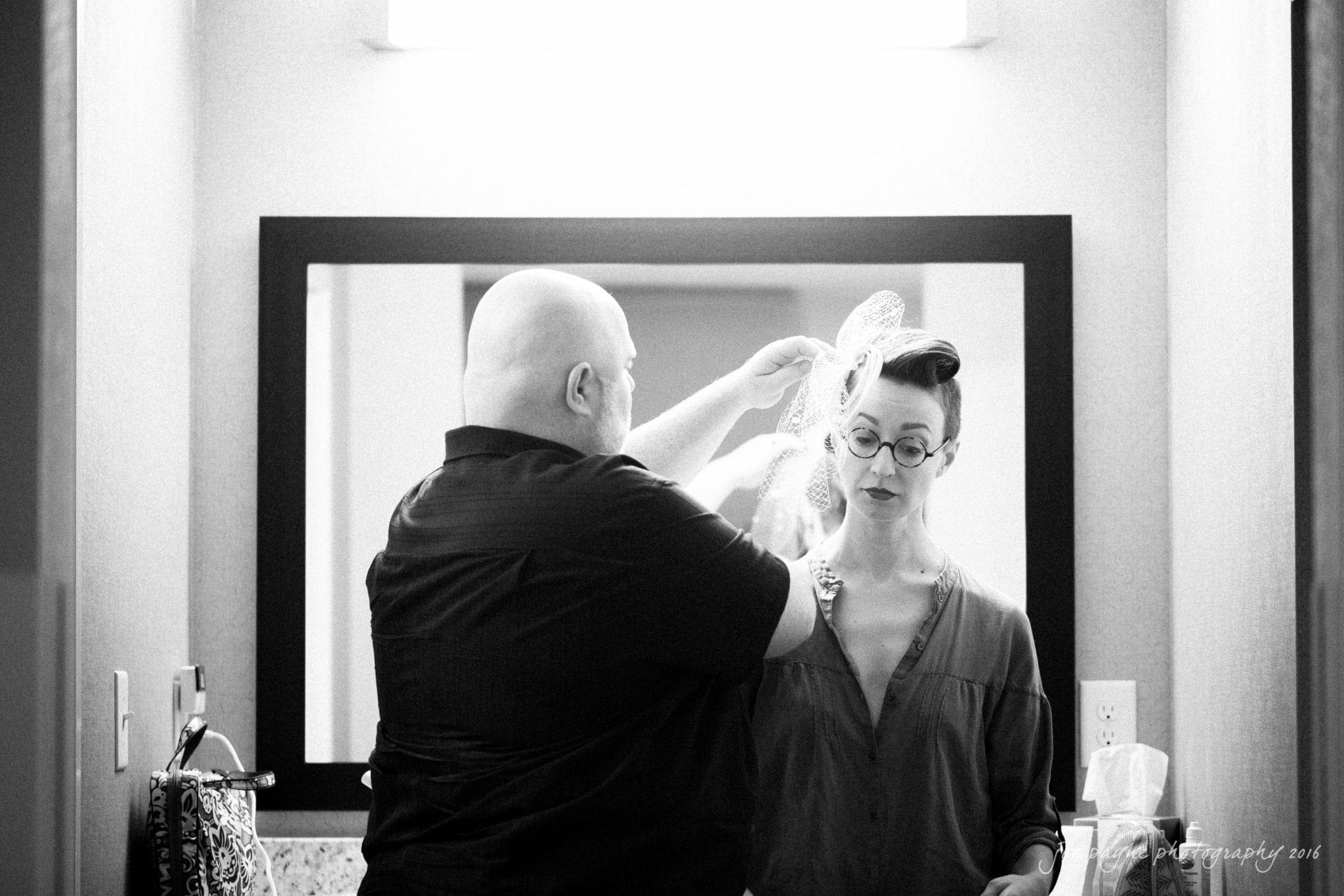 nc museum of art wedding photography - jade & frank A person helps another with a veil in a bathroom. The scene is in black and white, and they stand in front of a mirror with a counter in front of them. The person wearing the veil has short hair and glasses.