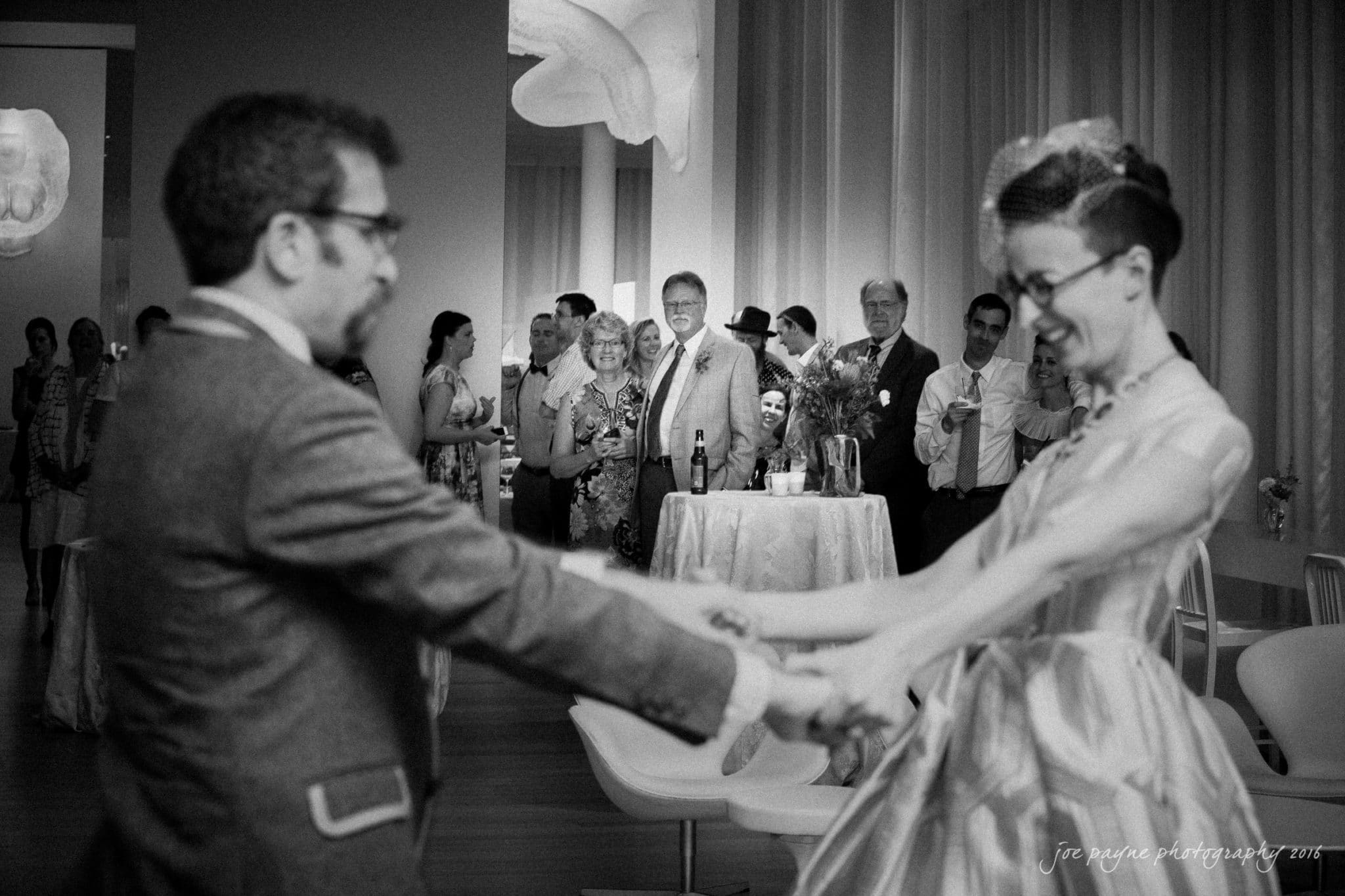 nc museum of art wedding photography - jade & frank A couple dances together at a wedding reception. The bride is in a white dress, and the groom wears a suit. Guests in the background watch and smile, with tables and drinks around them. The mood is joyful and celebratory.