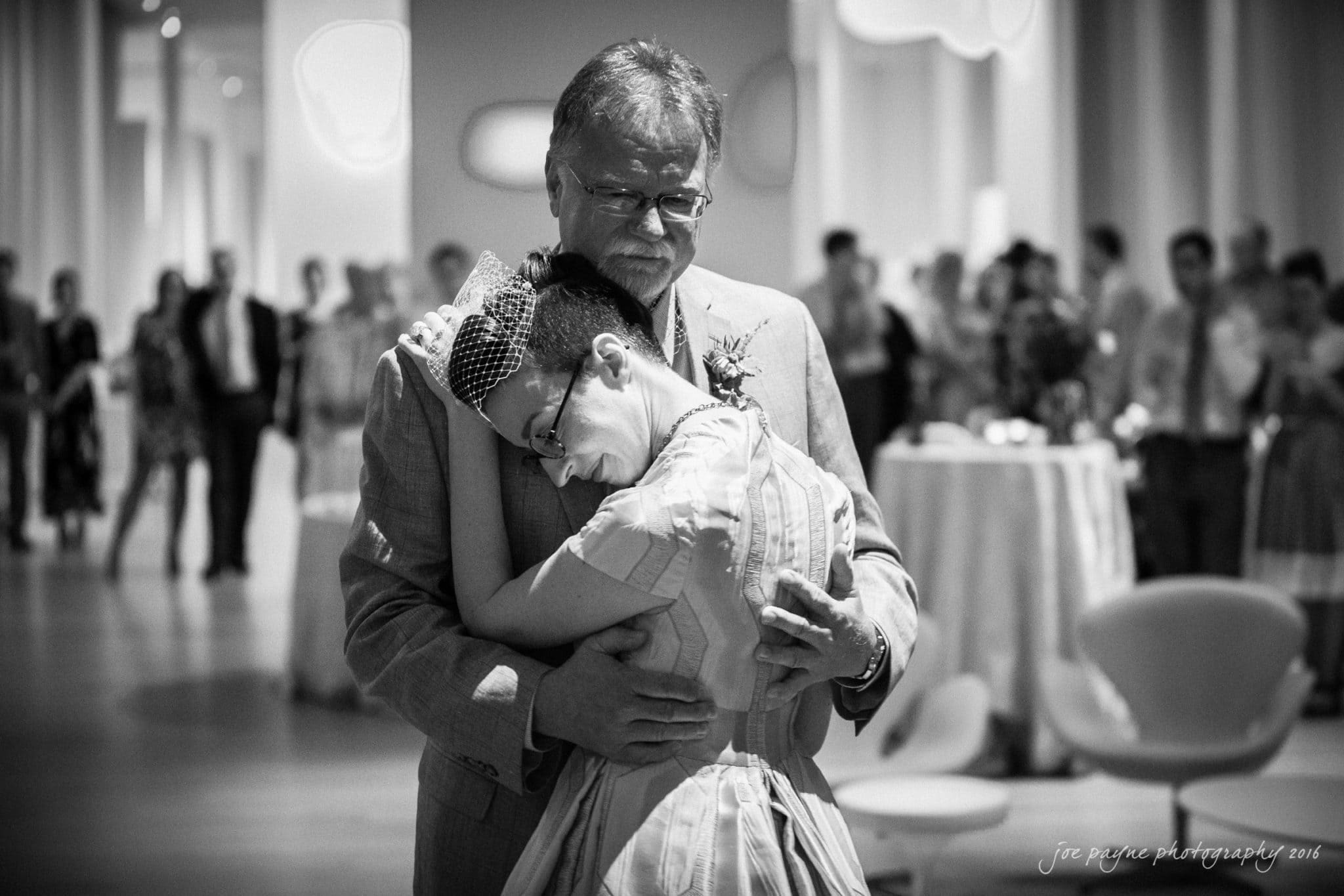 nc museum of art wedding photography - jade & frank A black-and-white photo of a bride in a vintage dress and birdcage veil hugging an older man in a suit, possibly her father. They stand closely, surrounded by blurred guests at a wedding reception. The atmosphere is emotional and tender.