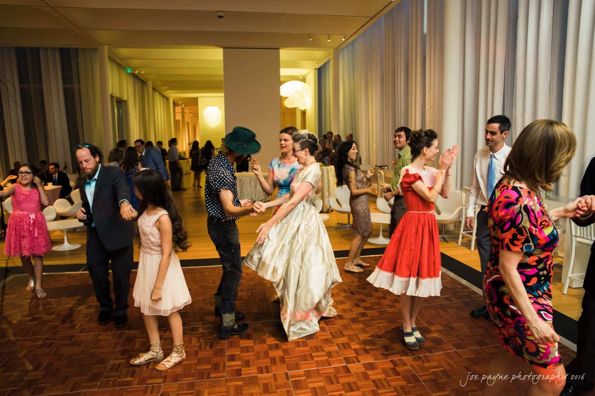 nc museum of art wedding photography - jade & frank A lively wedding reception scene with guests dancing energetically. The bride in a white dress and groom in a dark outfit are at the center, surrounded by others in colorful dresses and suits. The venue has soft lighting and elegant decor.