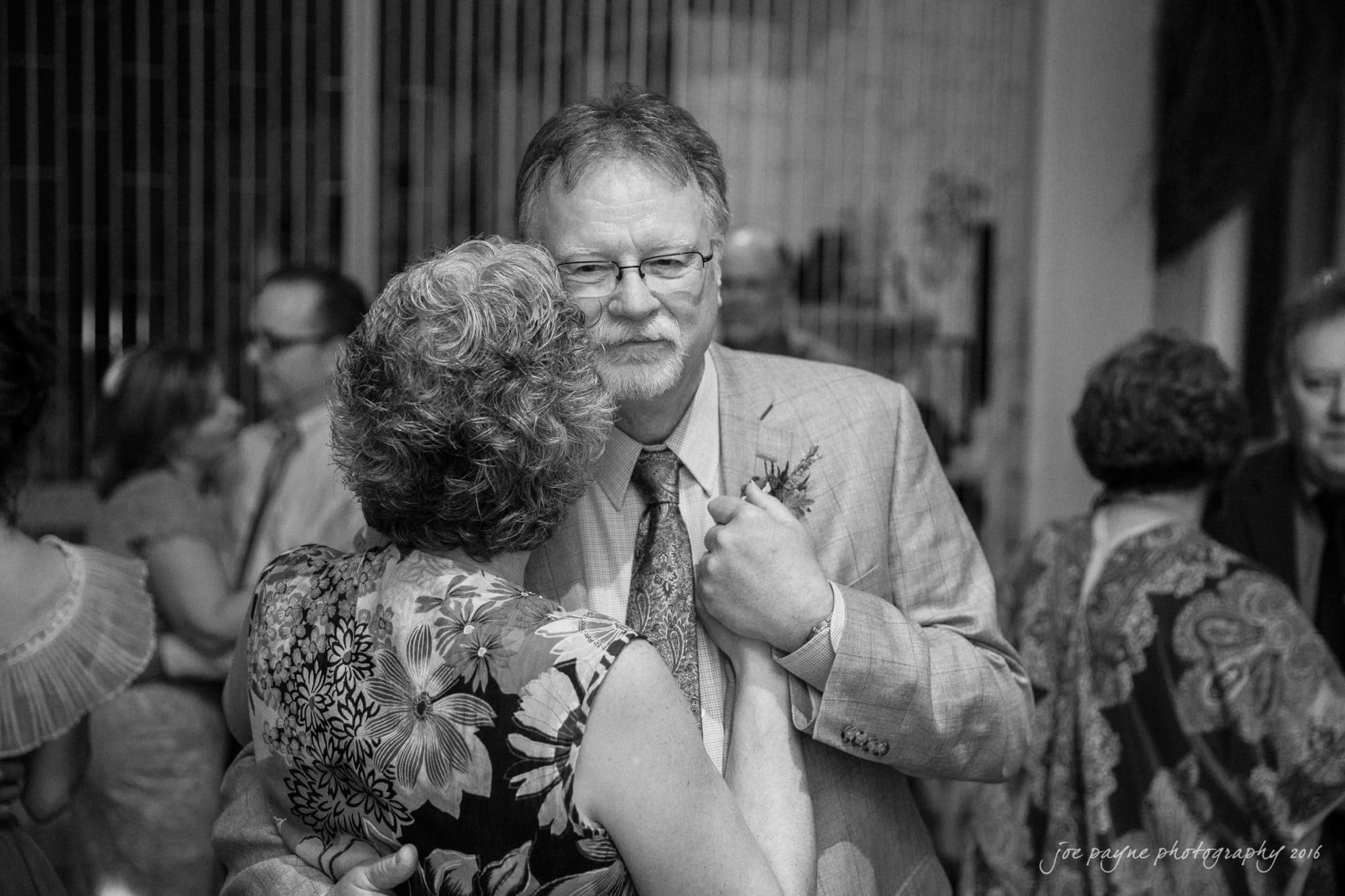nc museum of art wedding photography - jade & frank A man and a woman share a close embrace in a dimly lit room, surrounded by others in the background. The man wears glasses and a suit, while the woman has curly hair and a floral dress. The atmosphere is warm and intimate.