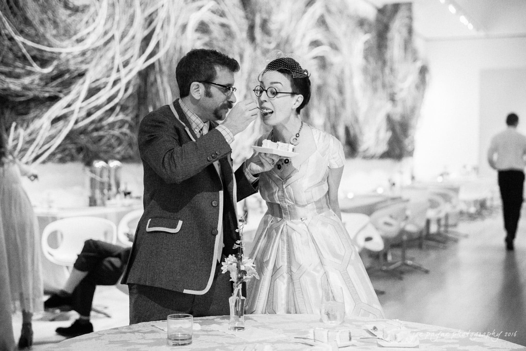 nc museum of art wedding photography - jade & frank A smiling couple stands next to a table in a stylish venue. The man, wearing a suit, appears to be feeding cake to the woman in a vintage-style dress. The background features abstract art and modern furniture.