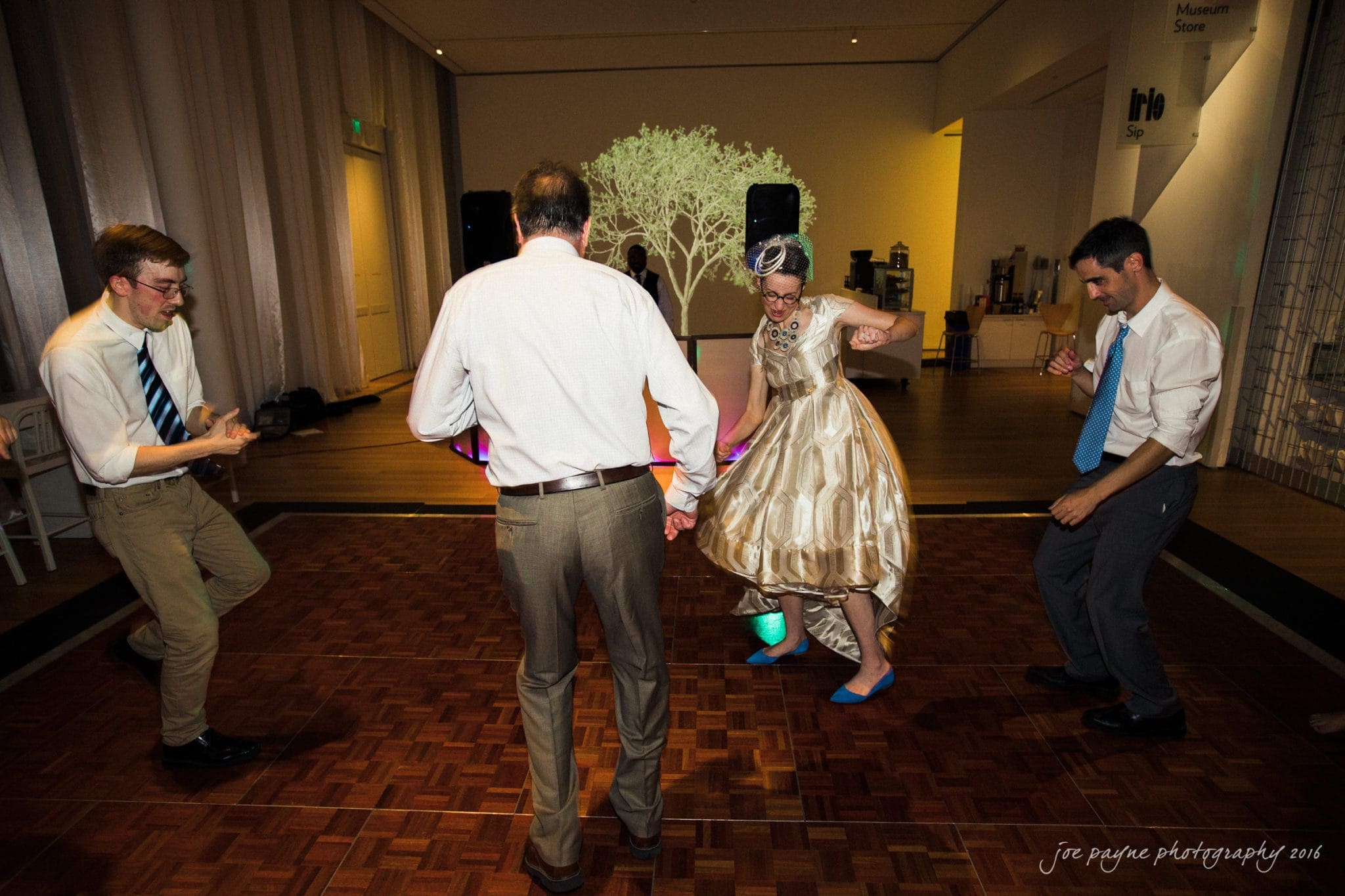 nc museum of art wedding photography - jade & frank A group of four people in formal attire are dancing energetically on a wooden dance floor. A projected image of a tree is visible on the wall in the background. The atmosphere is lively and joyful.