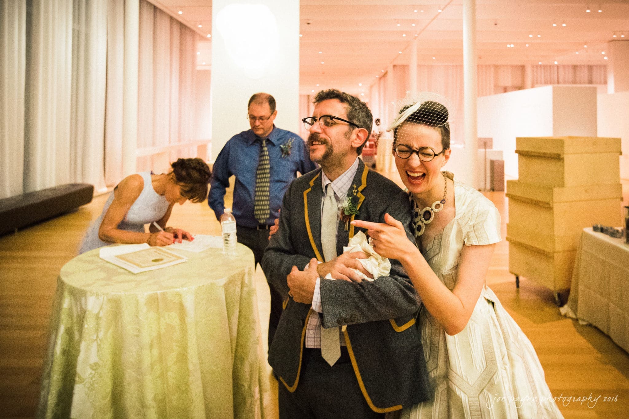 nc museum of art wedding photography - jade & frank A joyful bride in a vintage dress and veil laughs and points while standing beside a groom wearing a suit with yellow trim. Two guests are in the background; one is signing a document on a table. The scene is set in a bright, modern venue.