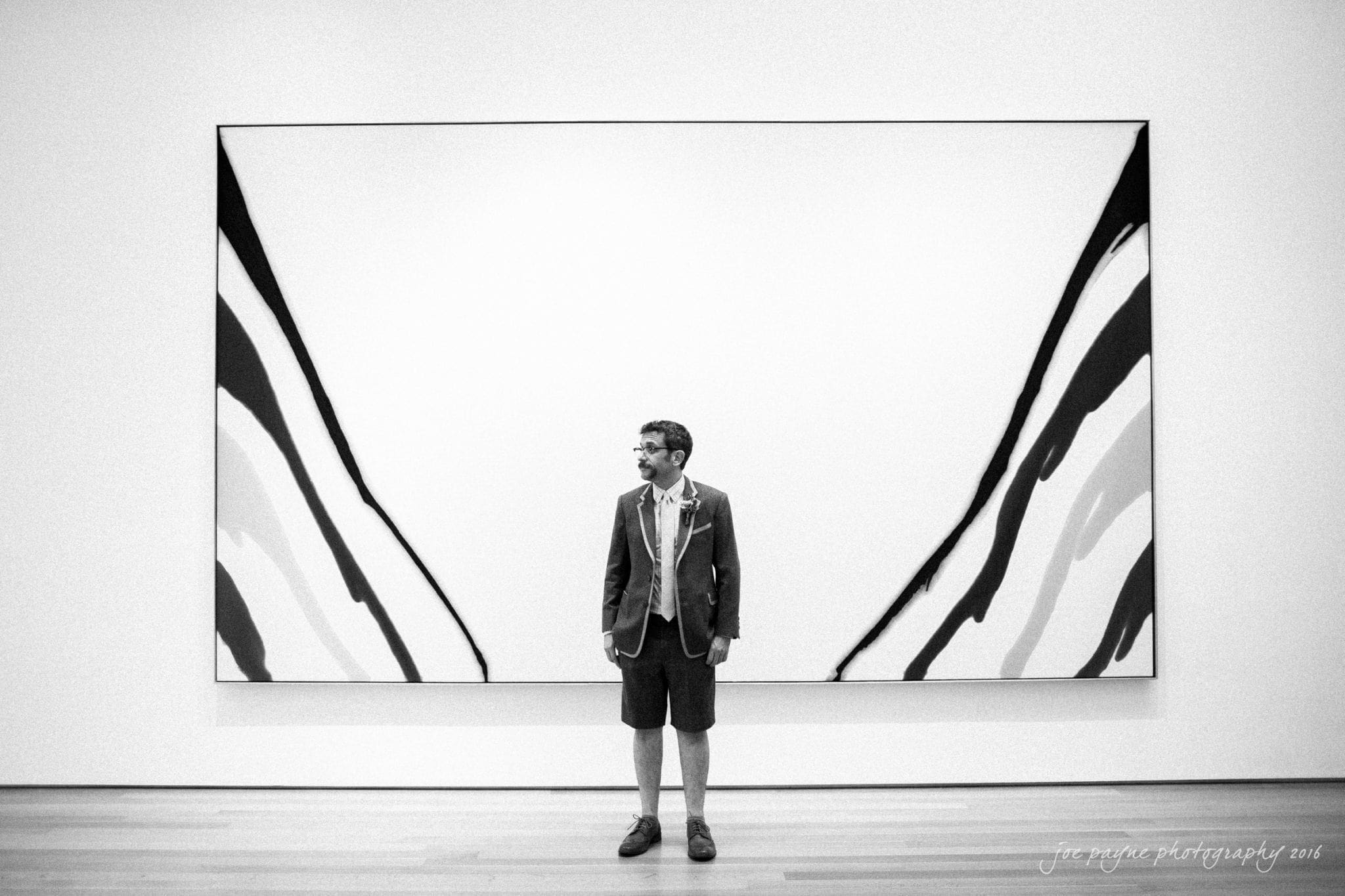 nc museum of art wedding photography - jade & frank A person in a suit and shorts stands in front of a large abstract black-and-white painting with bold, curving lines. The setting appears to be an art gallery. The photo is in black and white.