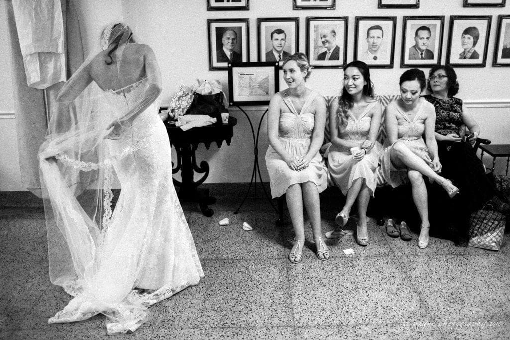 A bride adjusts her veil while four women sit nearby, three in matching dresses and one in a dark dress. The rooms walls are adorned with framed portraits. The scene captures a candid, pre-wedding moment.