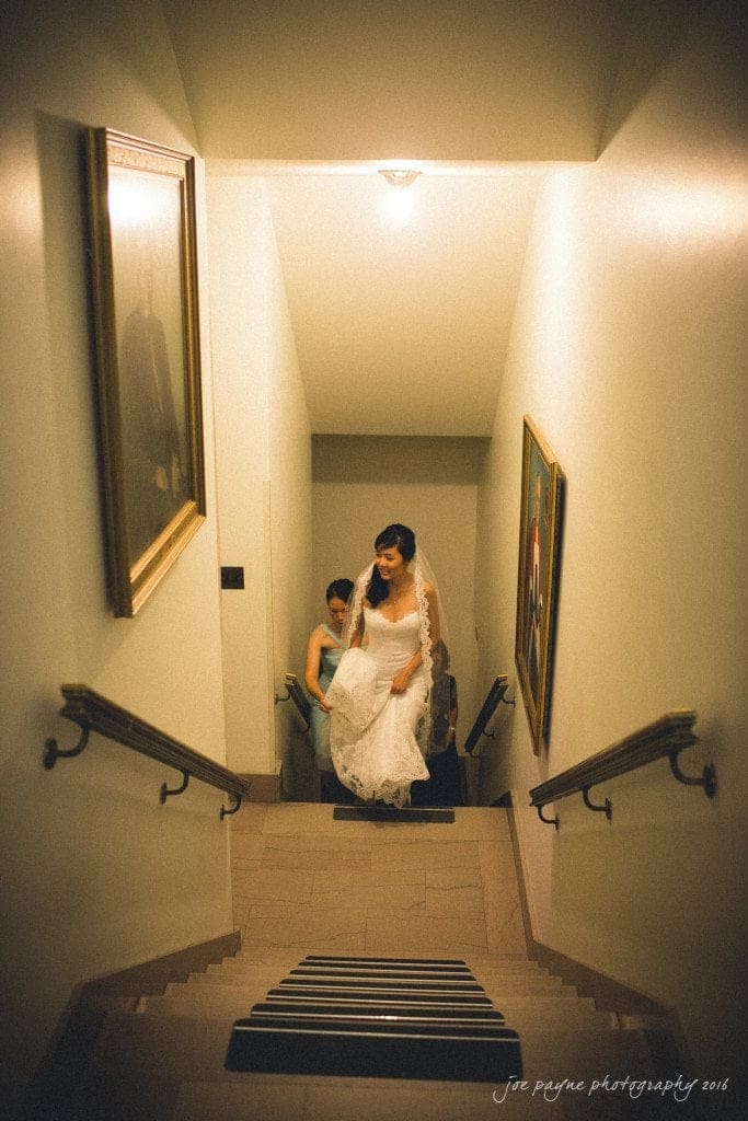 A bride in a white gown descends a dimly lit staircase, assisted by another woman in a blue dress. The walls are adorned with large portraits, and warm lighting casts soft shadows on the stairs.