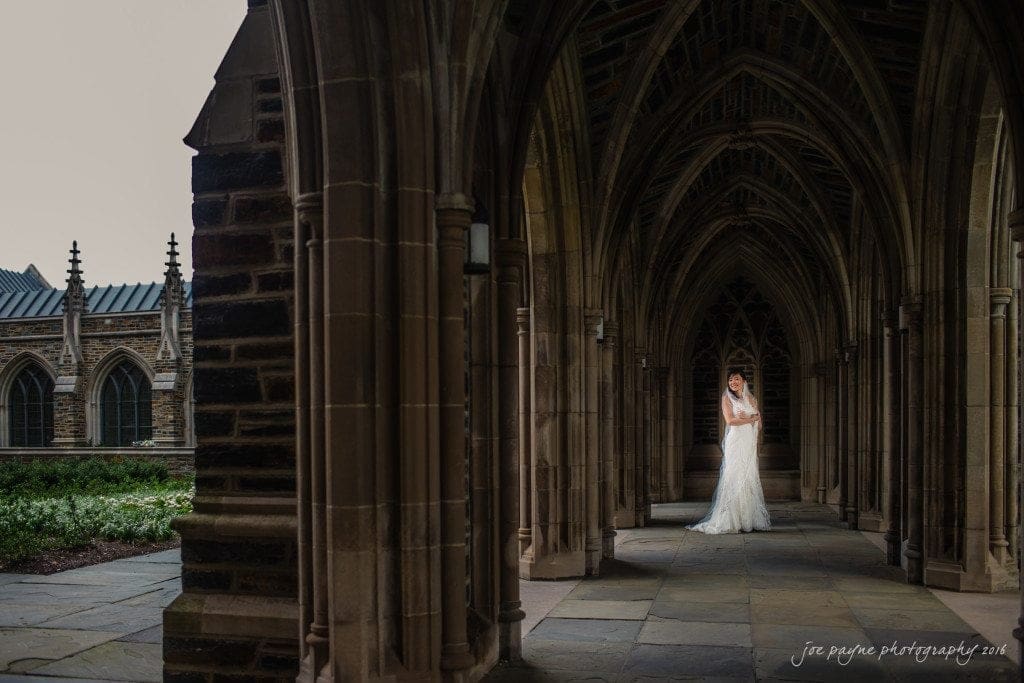 A bride in a white gown stands alone in an elegant, arched stone corridor. The gothic architecture frames her as soft light highlights the intricate design details.