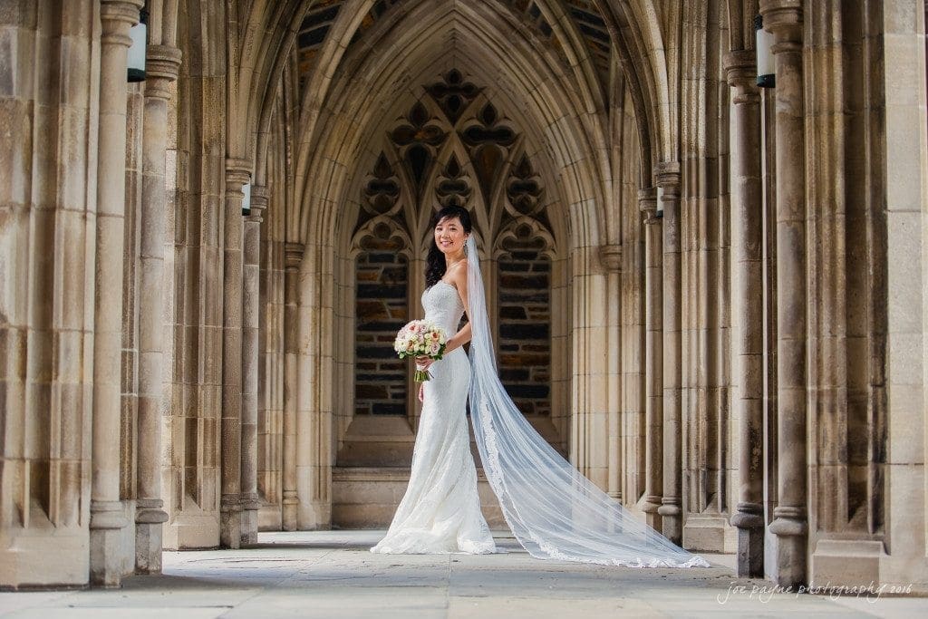A bride in a white gown with a long train stands in a stone archway, holding a bouquet. The intricate architecture surrounds her, creating a serene and elegant setting.