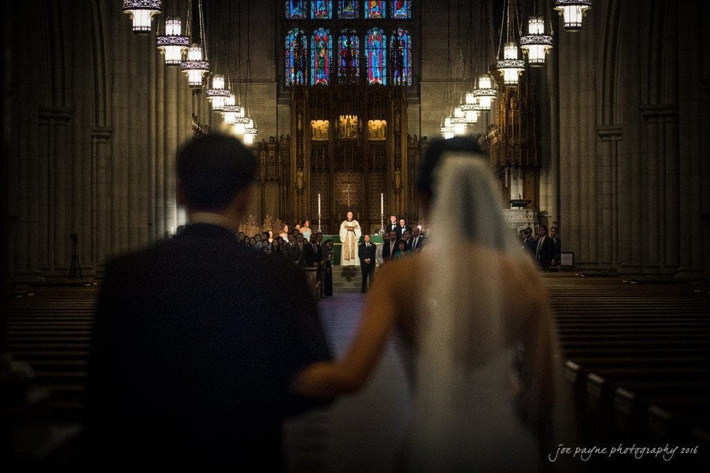 A bride and groom walk down the aisle of a grand cathedral, approaching a priest at the altar. The cathedral is adorned with ornate stained glass windows and hanging chandeliers, creating a solemn and beautiful atmosphere.