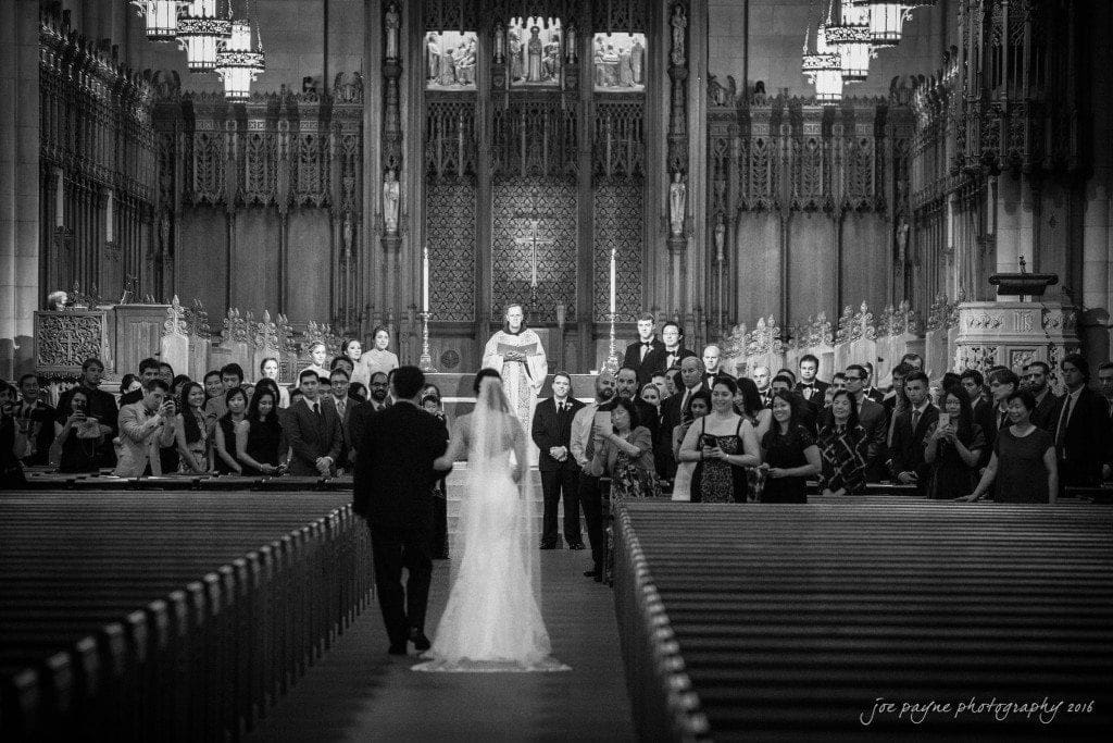 A black-and-white photo of a wedding ceremony inside a large, ornate church. The bride in a veil and the groom walk down the aisle, surrounded by seated guests. The priest stands in front of an elaborate altar. The image is signed Joe Payne Photography 2016 at the bottom.