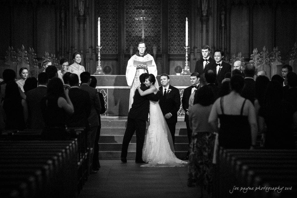 A bride and groom embrace in front of a priest at the altar during a wedding ceremony. Wedding party members stand nearby, and guests are seated in pews, watching the scene unfold. The setting is a large, ornate church.