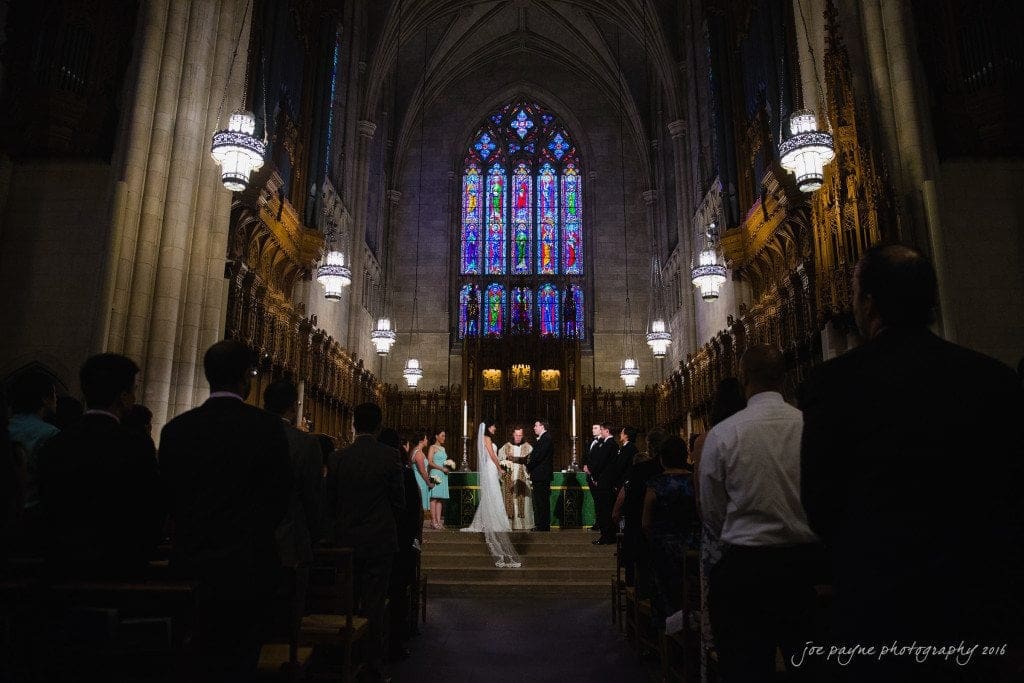 A couple stands at the altar in a grand cathedral during their wedding ceremony. The scene is illuminated by ornate chandeliers and features a large stained-glass window in the background, with guests seated on either side of the aisle.