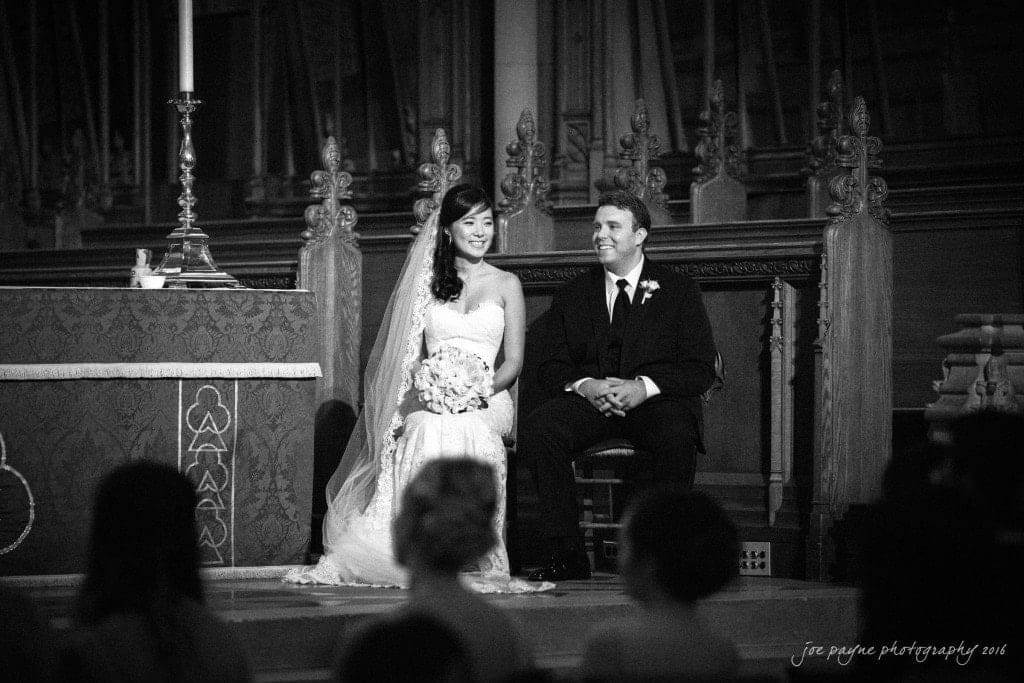 A bride and groom sit together in a church, smiling. The bride wears a white dress and holds a bouquet, while the groom wears a dark suit with a boutonniere. They are sitting near an altar, with candles and ornate woodwork visible in the background.