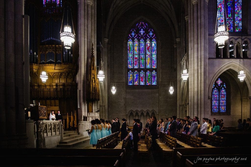 A church interior with high ceilings and stained glass windows. A wedding ceremony is taking place, with a bride and groom at the altar and people seated in pews. The setting is grand, featuring gothic-style architecture and soft lighting.