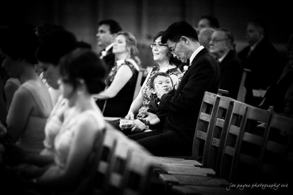 A black and white photo of wedding guests seated in a church. In focus, a man and woman with a small child on their laps look at the child. The scene captures a warm and intimate moment during the ceremony.