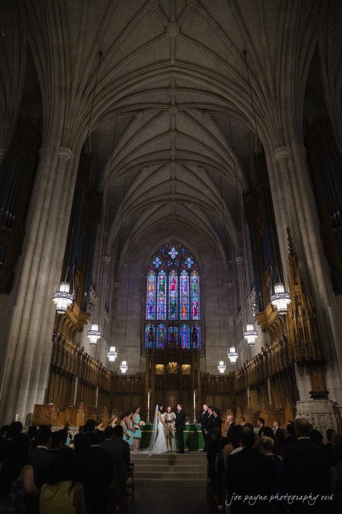 A couple stands at the altar in a grand cathedral, surrounded by wedding guests. The cathedral features tall arches, ornate woodwork, and colorful stained glass windows, creating a majestic and solemn atmosphere.