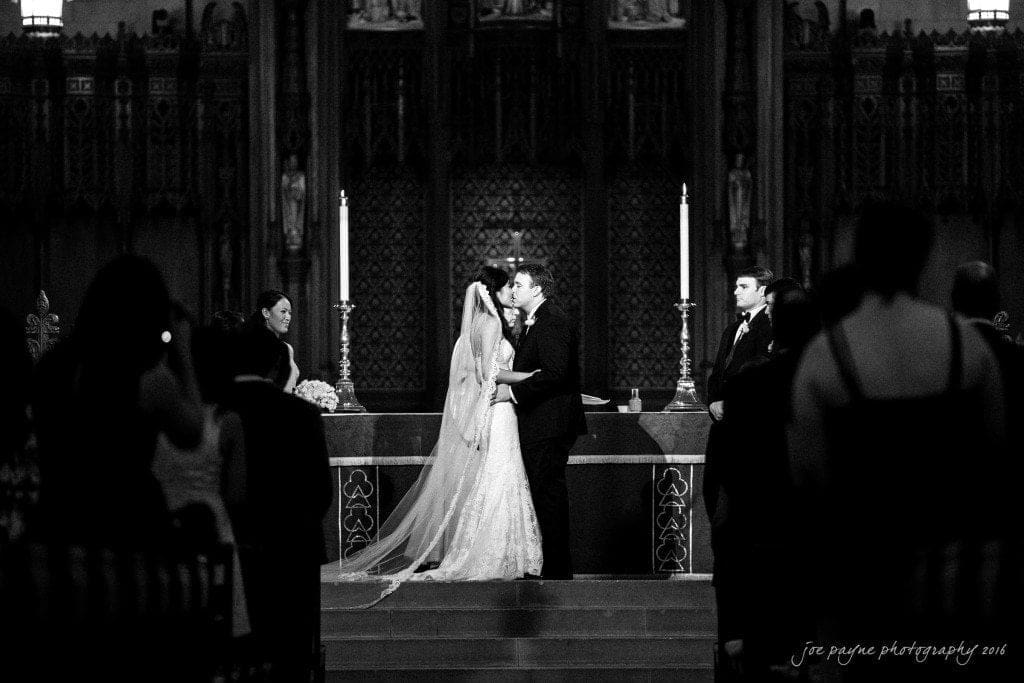 A bride and groom share a kiss at the altar in a church during their wedding ceremony. The scene is black and white, with attendants and guests witnessing the moment.