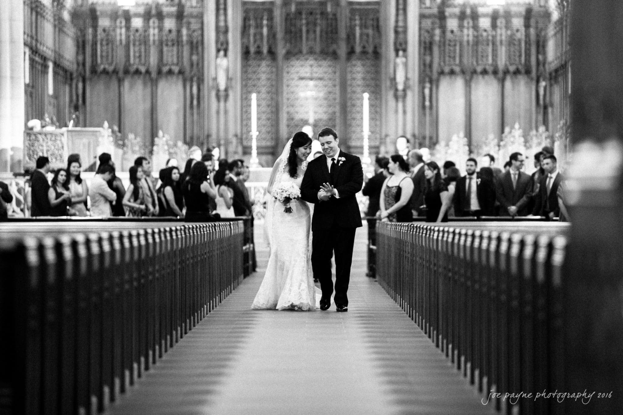 Duke Chapel Weddings A candid wedding recessional photograph at Duke Chapel, showing the groom happily admiring his new wedding ring as he walks arm-in-arm down the aisle with his bride.