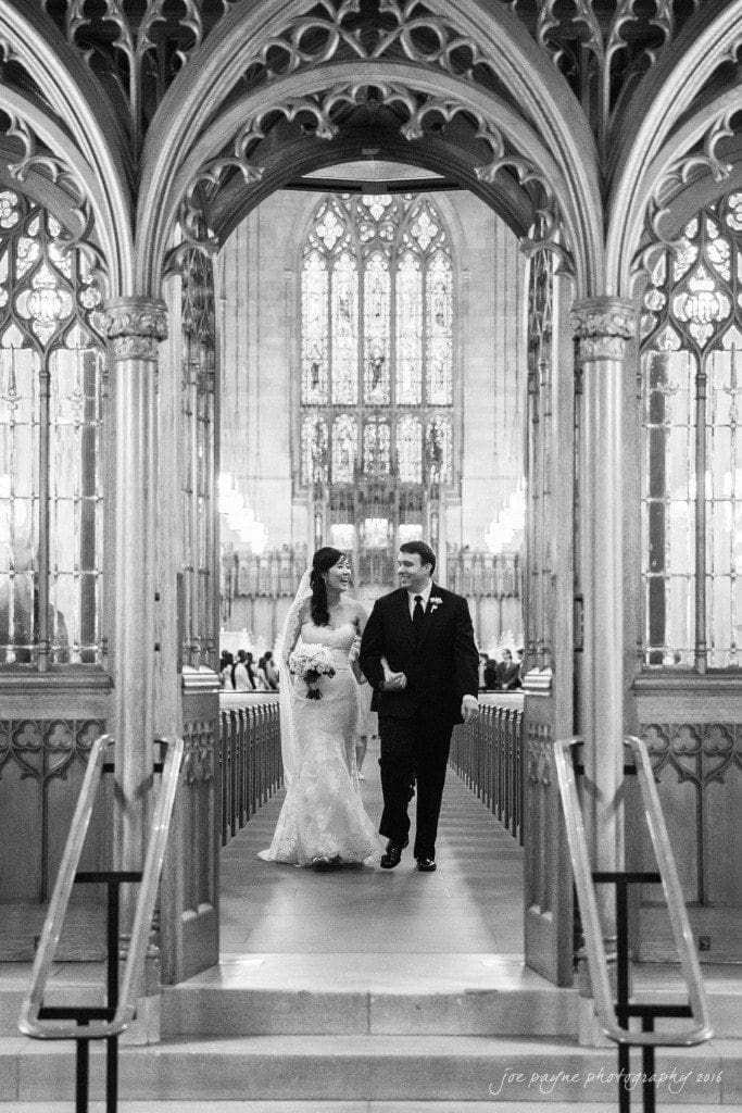 A bride and groom walk down the aisle of a grand, ornate church with intricate wooden carvings and large stained glass windows. The couple is holding hands and smiling, surrounded by elegant architectural details.