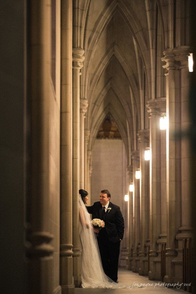 A couple stands intimately within a grand, arched hallway adorned with soft lighting. The bride wears a long, flowing veil and holds a bouquet, while the groom, in a dark suit, gazes at her lovingly. The atmosphere is romantic and serene.