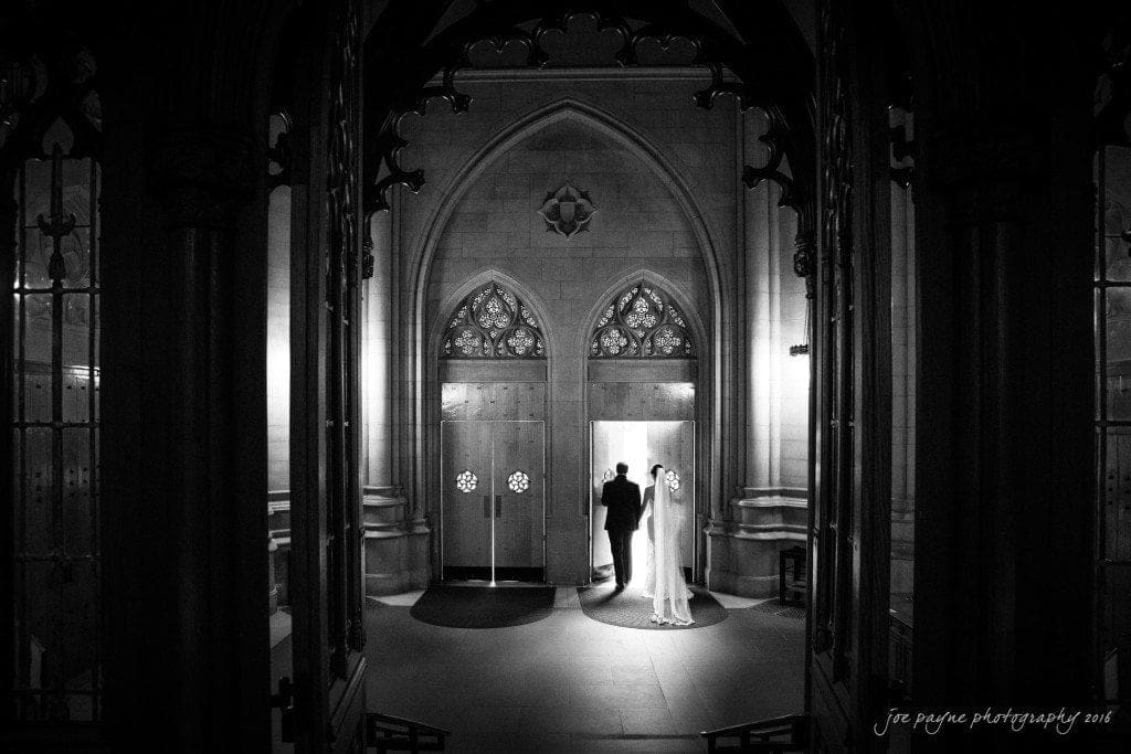 A bride and groom holding hands stand in the grand entrance of a cathedral. The dramatic black and white setting highlights the arched doors and ornate architecture, creating a timeless and romantic scene.