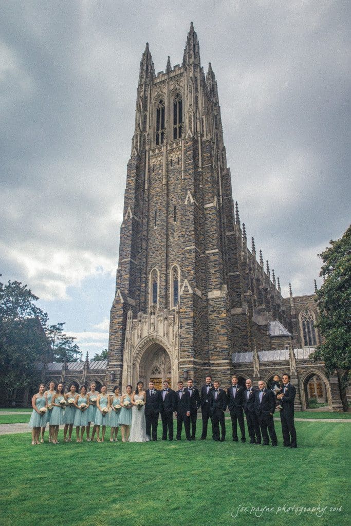 A wedding party poses in front of a large, gothic-style cathedral under a cloudy sky. The party includes bridesmaids in pale dresses and groomsmen in dark suits standing on a lush green lawn.
