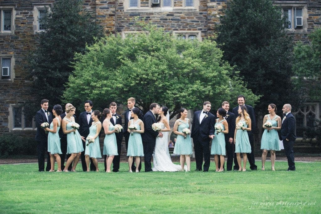 A wedding party stands on a green lawn in front of a stone building. The bridesmaids wear light teal dresses, and the groomsmen wear dark suits. The bride and groom are in the center, surrounded by their wedding party holding bouquets.