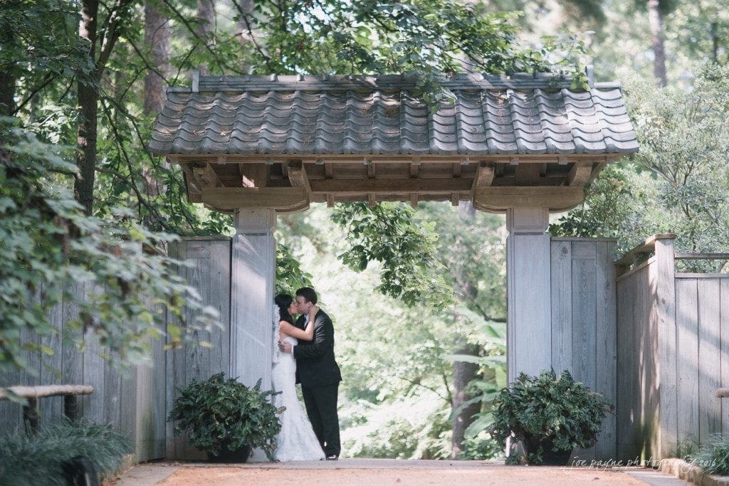 A bride and groom embrace under a wooden gate in a lush garden. The bride wears a white dress, and the groom is in a black suit. Sunlight filters through the trees, creating a serene, romantic atmosphere.