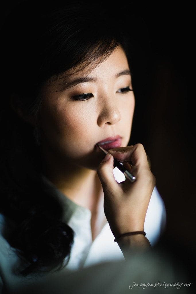 Close-up of a person with long dark hair having their makeup applied, focusing on their lips, against a dark background. The lighting highlights their face, emphasizing the makeup detail.