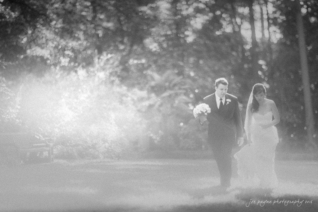 A black-and-white photo of a bride and groom walking through a sunlit, wooded area. The bride holds a bouquet and the groom wears a suit. Sunlight filters through the trees, creating a soft, dreamy atmosphere.