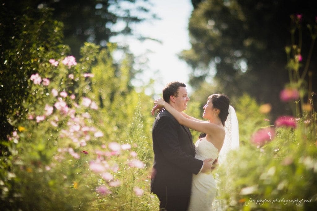A bride and groom gaze at each other lovingly in a sunlit garden surrounded by pink and purple wildflowers. The bride wears a white dress with a veil, and the groom is in a black suit. The scene is serene and romantic.