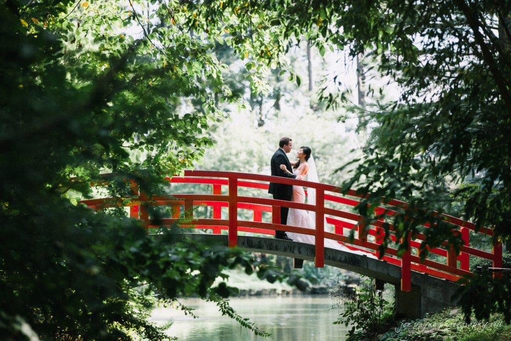 A couple stands on a red wooden bridge surrounded by lush greenery. The bride is in a white dress, and the groom is in a dark suit. Sunlight filters through the trees, creating a serene and romantic atmosphere.