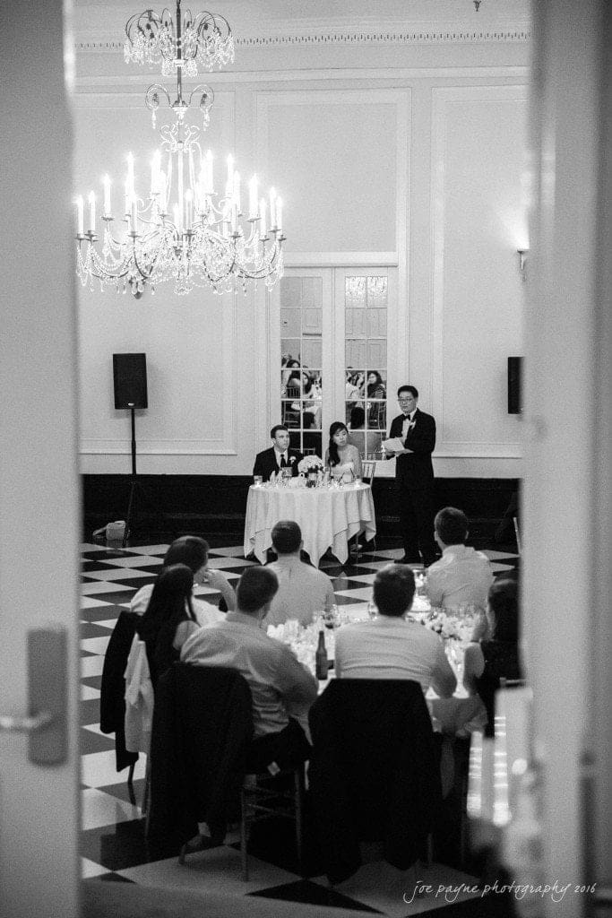 A black and white photo of a wedding reception in an elegant ballroom. Guests are seated at round tables with a checkered floor. A person stands giving a speech near a couple seated at a small table. Chandeliers hang from the ceiling.