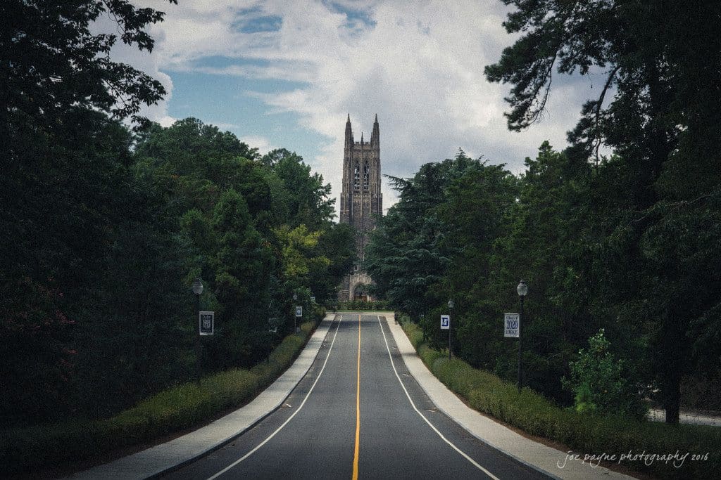 A long, straight road lined with trees leads to a tall, gothic-style tower in the distance. The sky is partly cloudy, and the scene has a serene, atmospheric quality.
