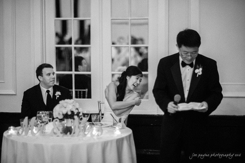 A black and white photo of a wedding scene. A man in a tuxedo stands holding a microphone and reading from a paper. A woman in a white gown is laughing and pointing her thumbs towards a seated man in a suit at a table with floral decorations.